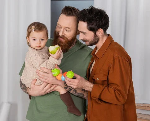 A toddler is held by a man with a beard, while another man in a brown shirt plays with colorful toys. The background is softly lit.