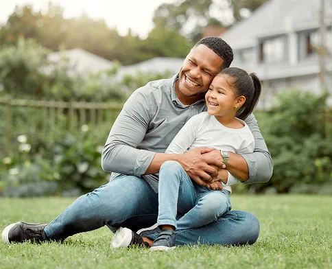 A parent and child sit together on the grass, enjoying a sunny day in a garden, with a cozy and affectionate pose.