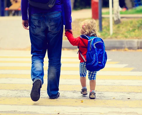 A parent and child cross a street together, holding hands. The child wears a red jacket and blue backpack, enjoying the moment.