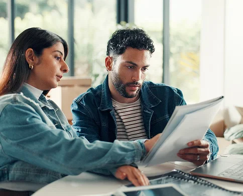 Two individuals collaborate at a table, reviewing documents, with a laptop and tablet nearby, in a bright, modern workspace.