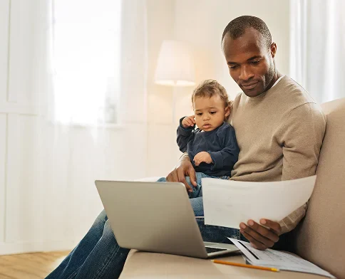 A person sits on a sofa with a laptop and papers, holding a child in their lap, in a bright, cozy room.