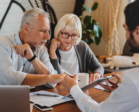 A man and woman discuss documents and plans with a professional at a table, surrounded by a laptop and office supplies.