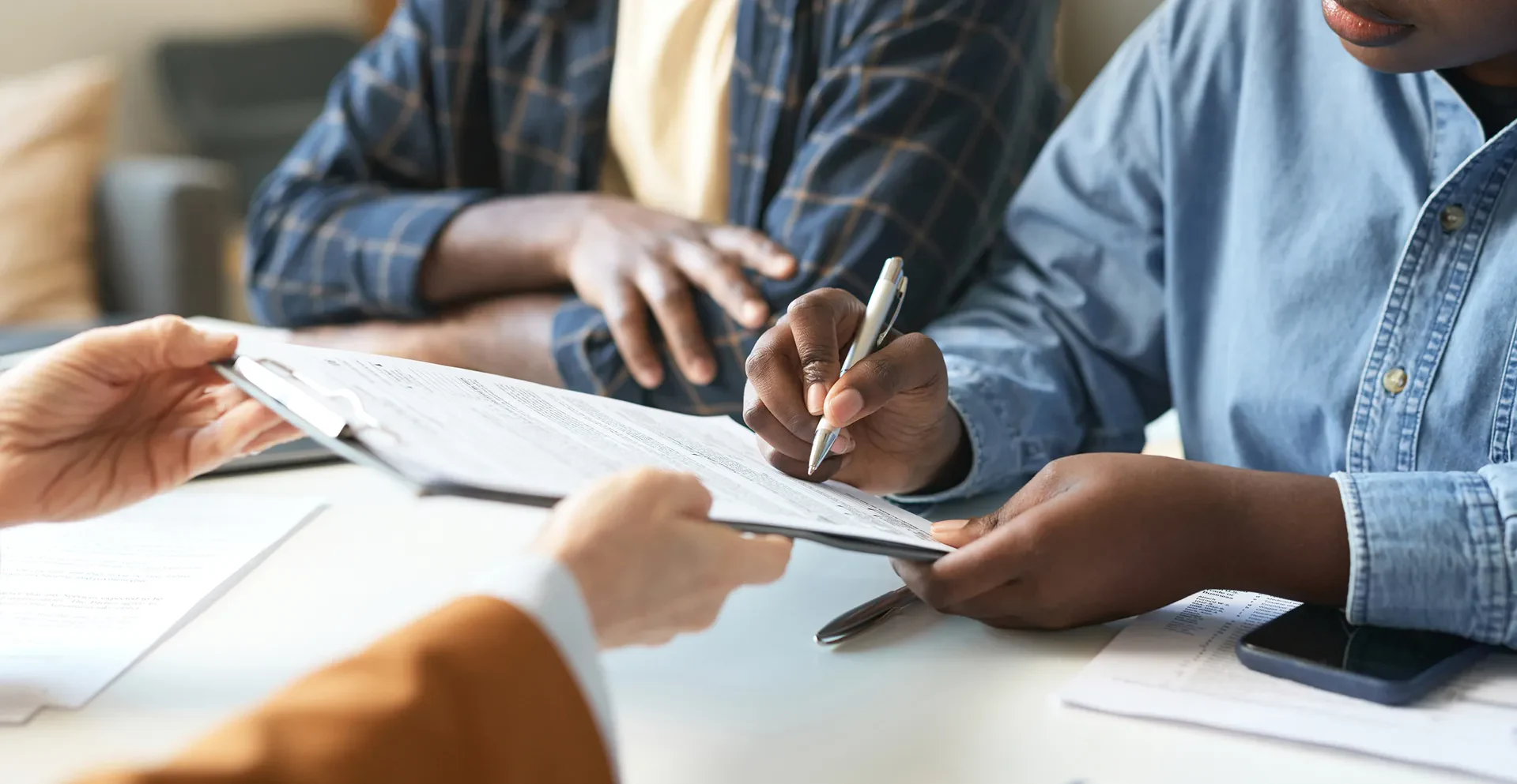 A close-up of hands exchanging a clipboard filled with documents. One person is signing while others observe attentively.